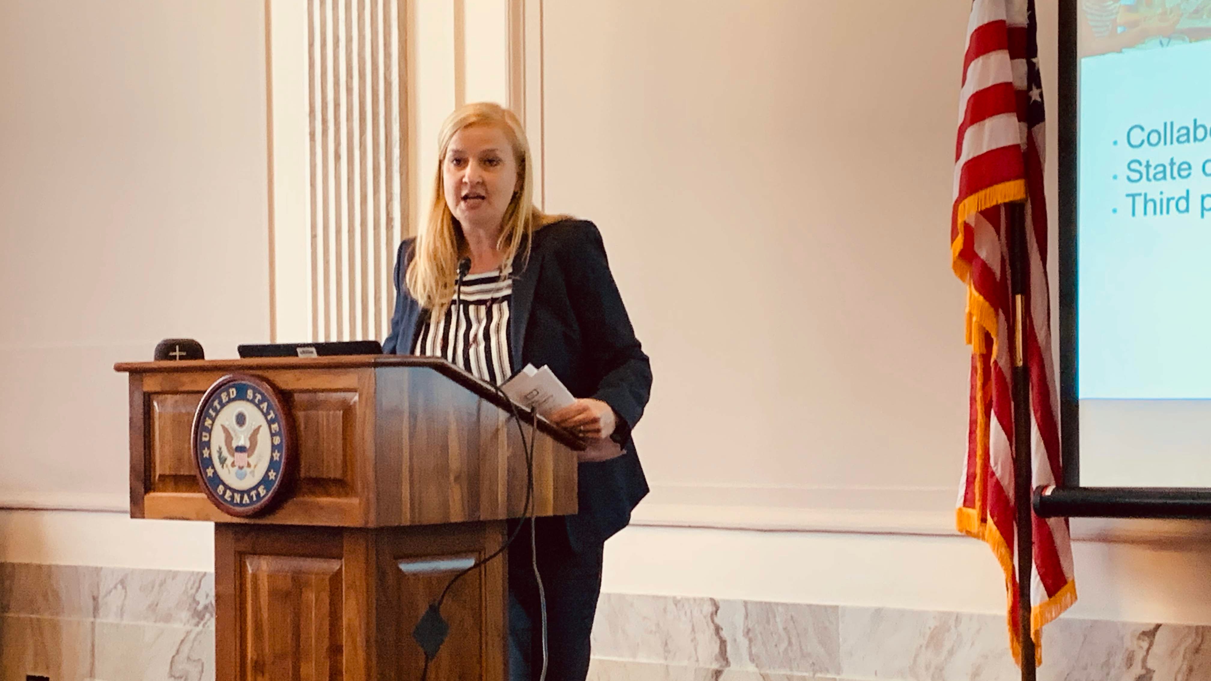 Christine Fox, presenting at podium bearing US Senate seal